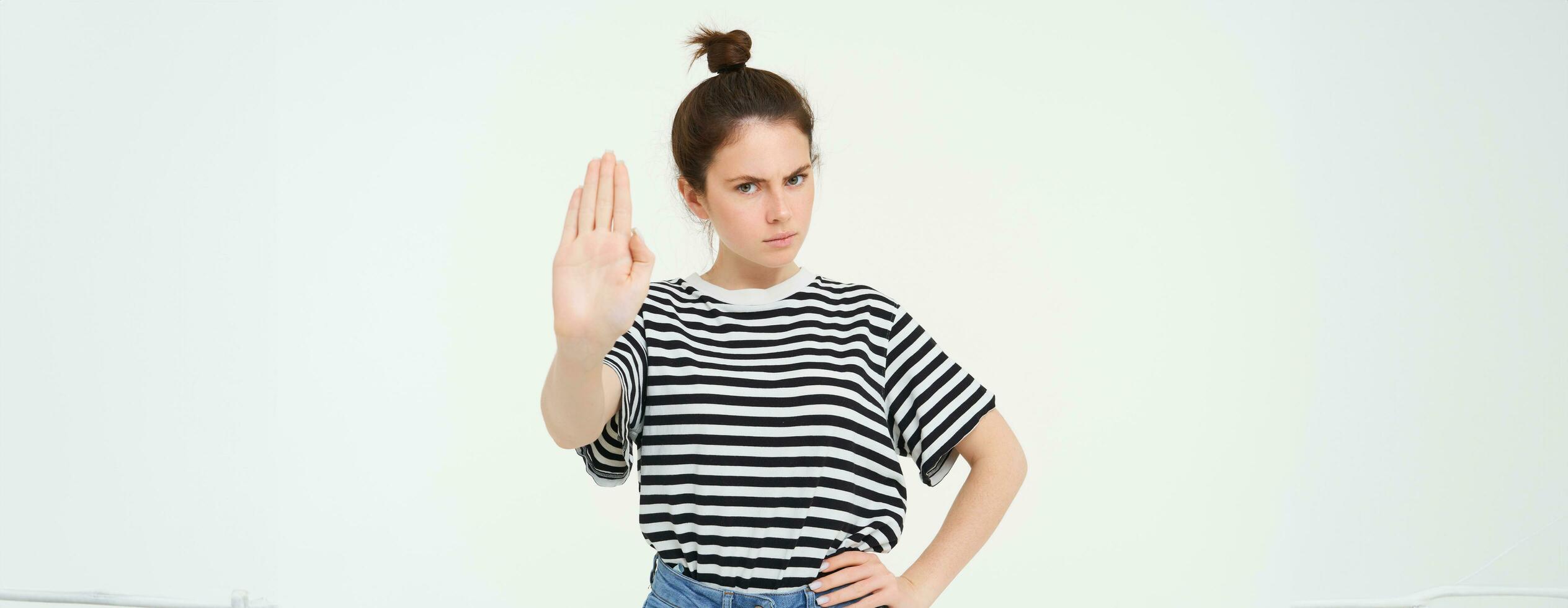 Image of beautiful woman with serious face, showing stop sign, extends one palm and frowns, stands over white background photo