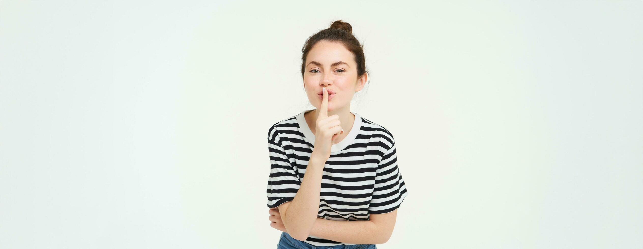 Portrait of cute girl keeps a secret, shushes, shhh gesture, presses finger to lips, gossiping, stands over white background photo