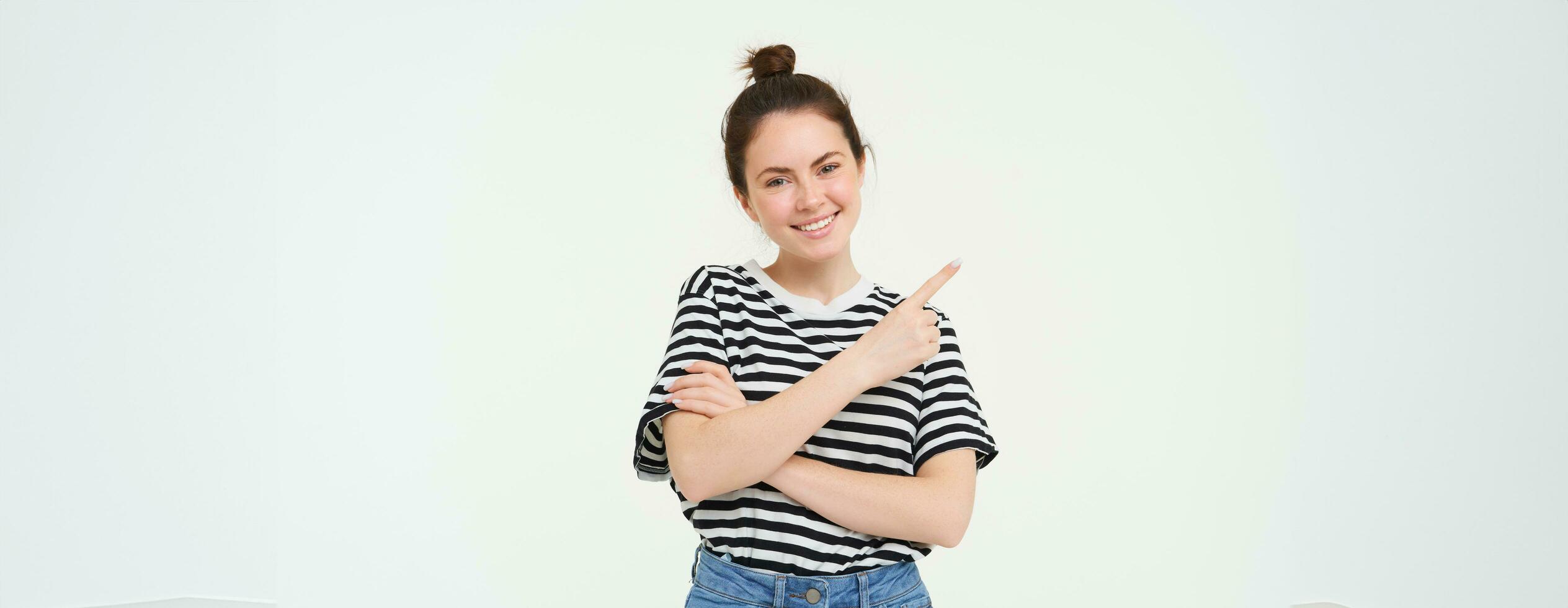 Portrait of cute smiling female model, wearing casual clothes, pointing finger left, showing advertisement, white background photo