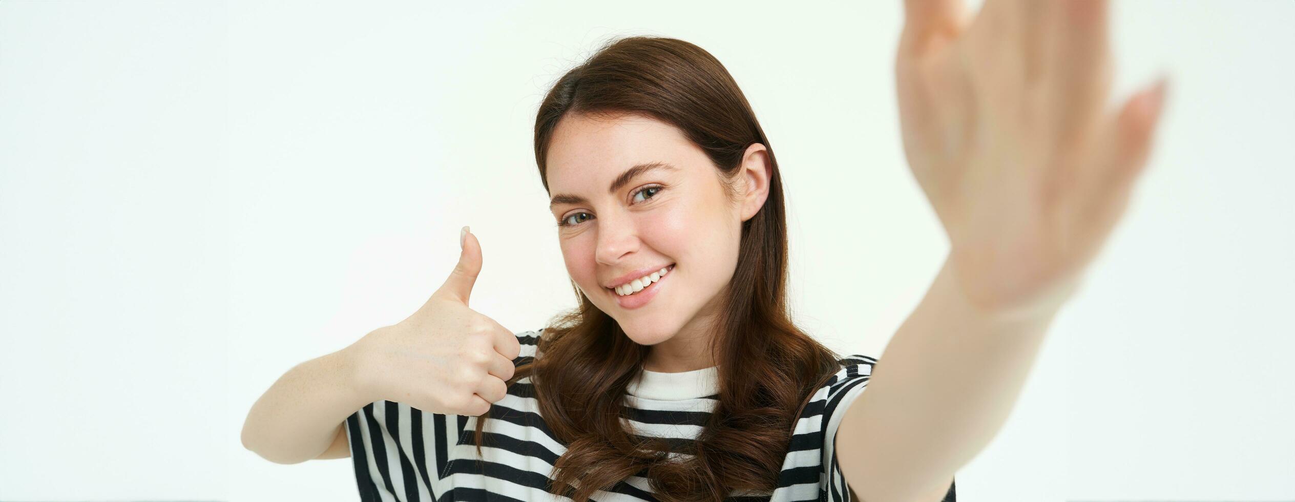Portrait of girl takes selfie with thumbs up next to something she recommends, likes and approves, stands over white background photo