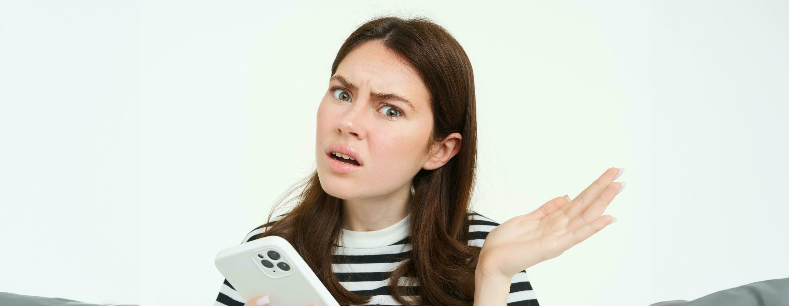 Portrait of woman shrugging, holding smartphone and looking confused at camera, isolated on white background photo