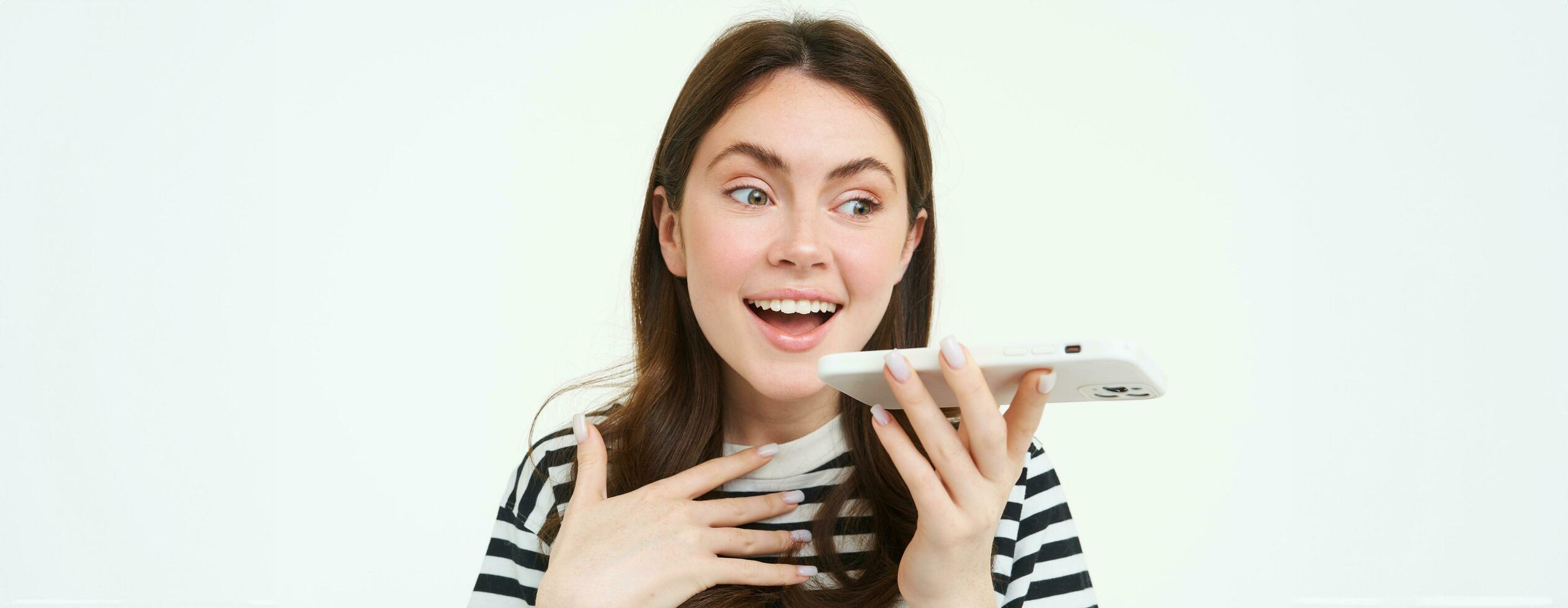 Image of cute brunette woman talking into speakerphone, holding mobile phone near mouth, records her voice, sends a voicemessage, using online translator app, white background photo