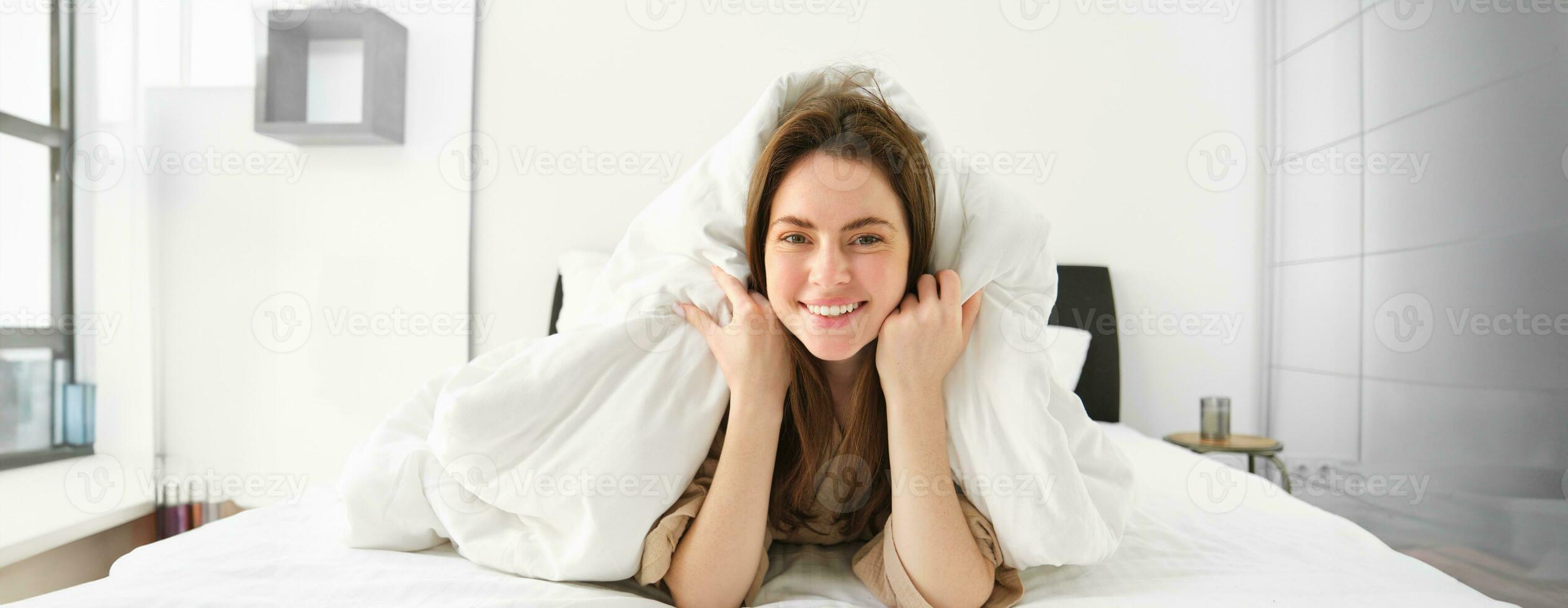 Cute girl with messy hair, lying in bed covered in white sheets duvet, smiling and laughing coquettish, spending time in her bedroom photo