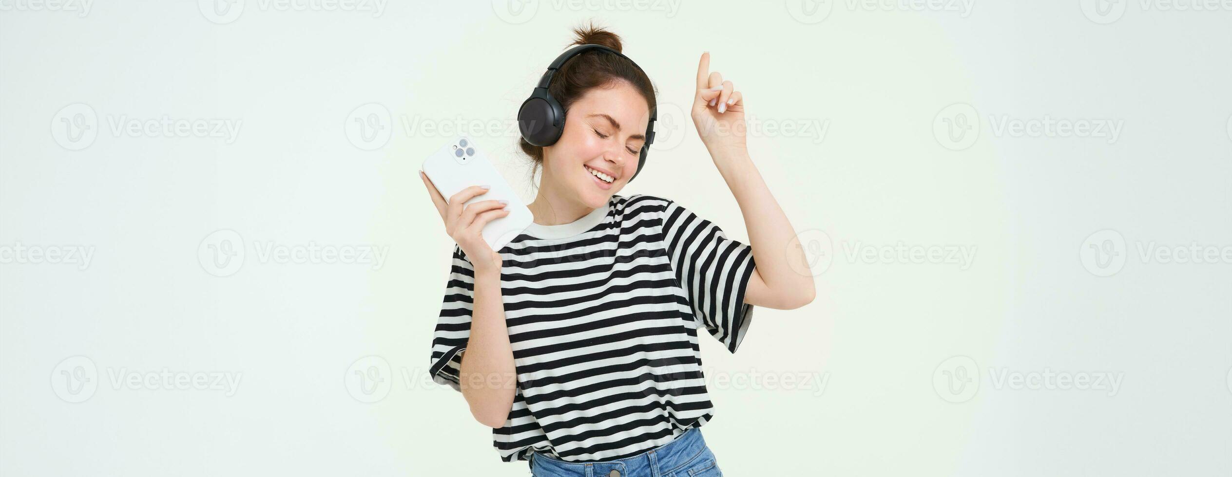 Portrait of happy woman with smartphone changes song on mobile phone streaming app, listens music in headphones, dancing against white background photo