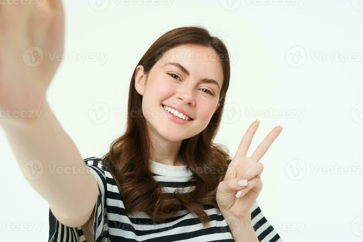 teléfono inteligente vista, mujer participación móvil teléfono cámara y tomando selfie con paz, signo v gesto, sonriente a cámara, posando terminado blanco antecedentes foto