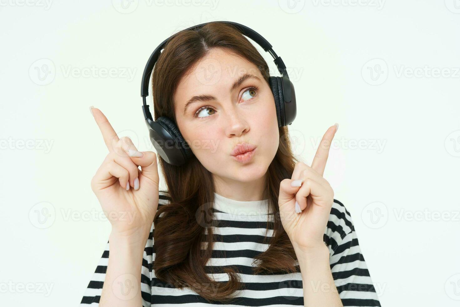 retrato de joven morena mujer en auriculares, escuchando música en auriculares, señalando dedos arriba, demostración bandera, anuncio publicitario, aislado terminado blanco antecedentes foto