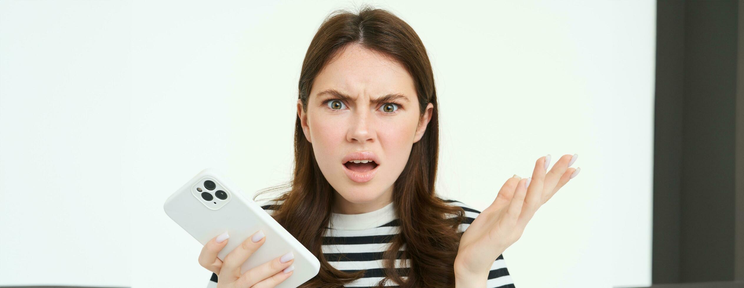 Portrait of angry, confused young woman shrugging shoulders while using mobile phone, holding smartphone with annoyed face expression, frowning, standing over white background photo