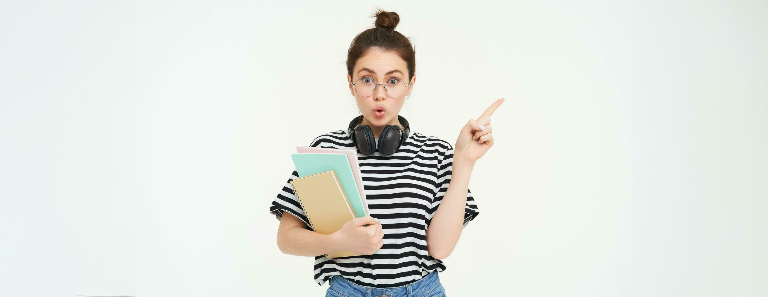 Portrait of girl with surprised face, interested in advertisement on the left, pointing at banner with curious face, wearing glasses, holding documents and notebooks in hand, white background photo