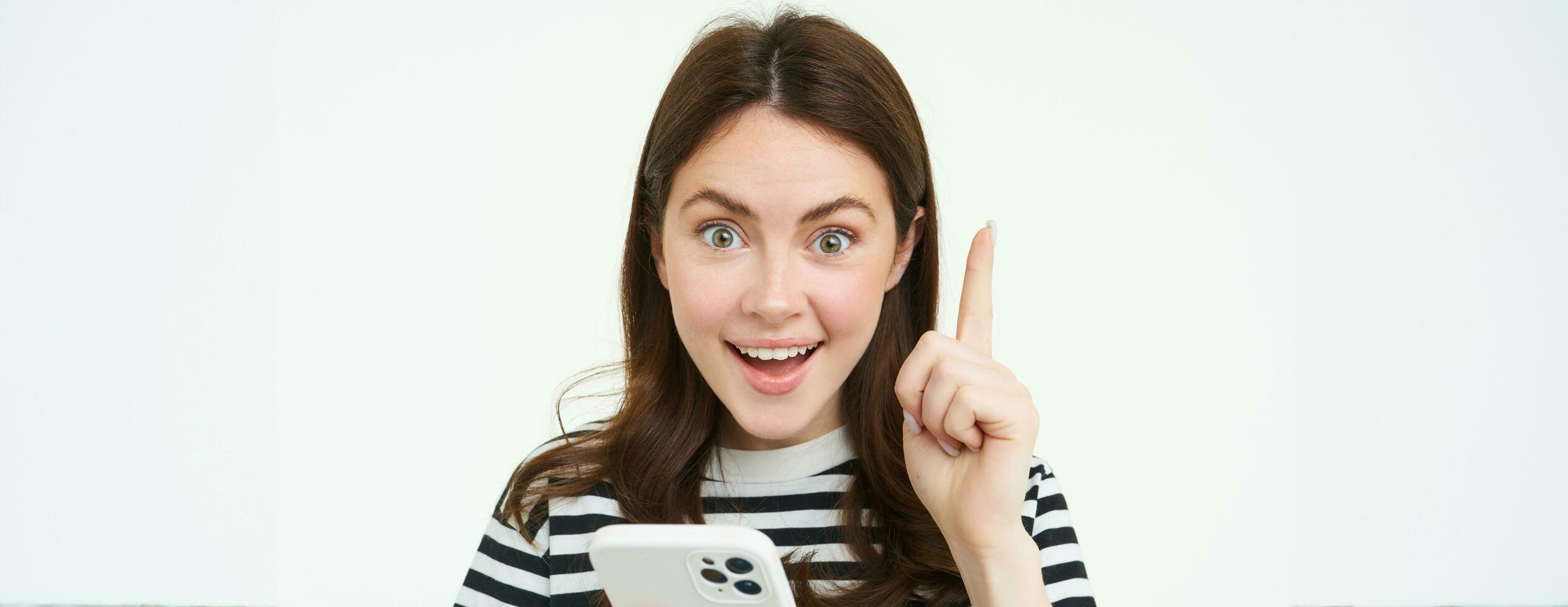 Got an idea. Smiling woman, raises her finger and holds smartphone, suggests something, makes eureka gesture, has a solution, white background photo