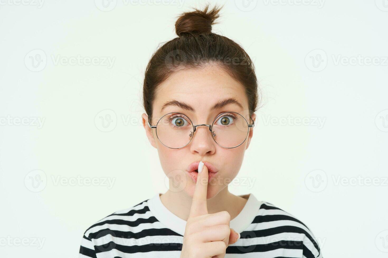 Portrait of girl with glasses, looks surprised, hughing, press finger to lips, shush shh gesture, asks to keep quiet, stands over white background photo