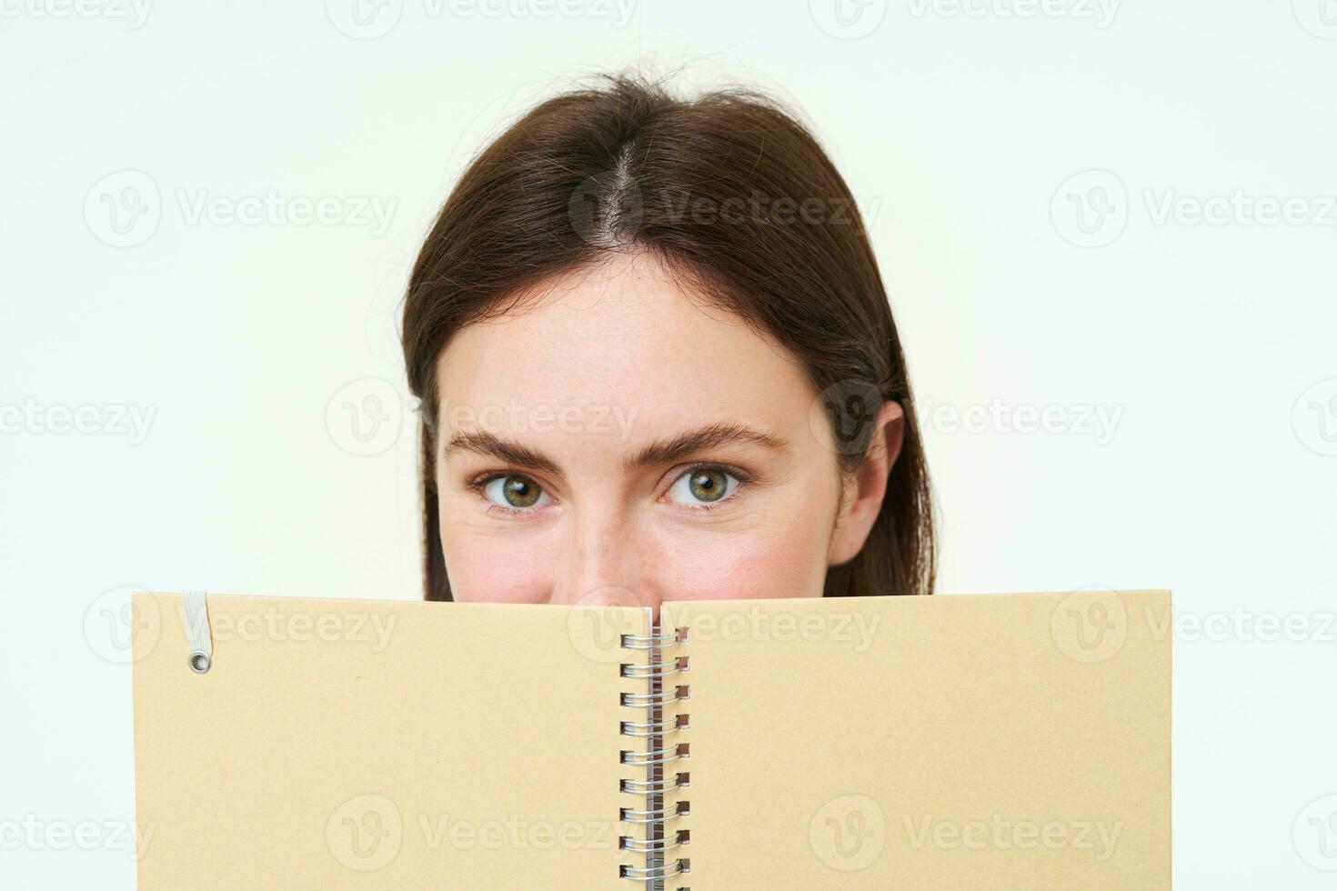Young woman holds notebook daily planner next to her face, writing down homework, making notes, looking thoughtful, standing over white background photo