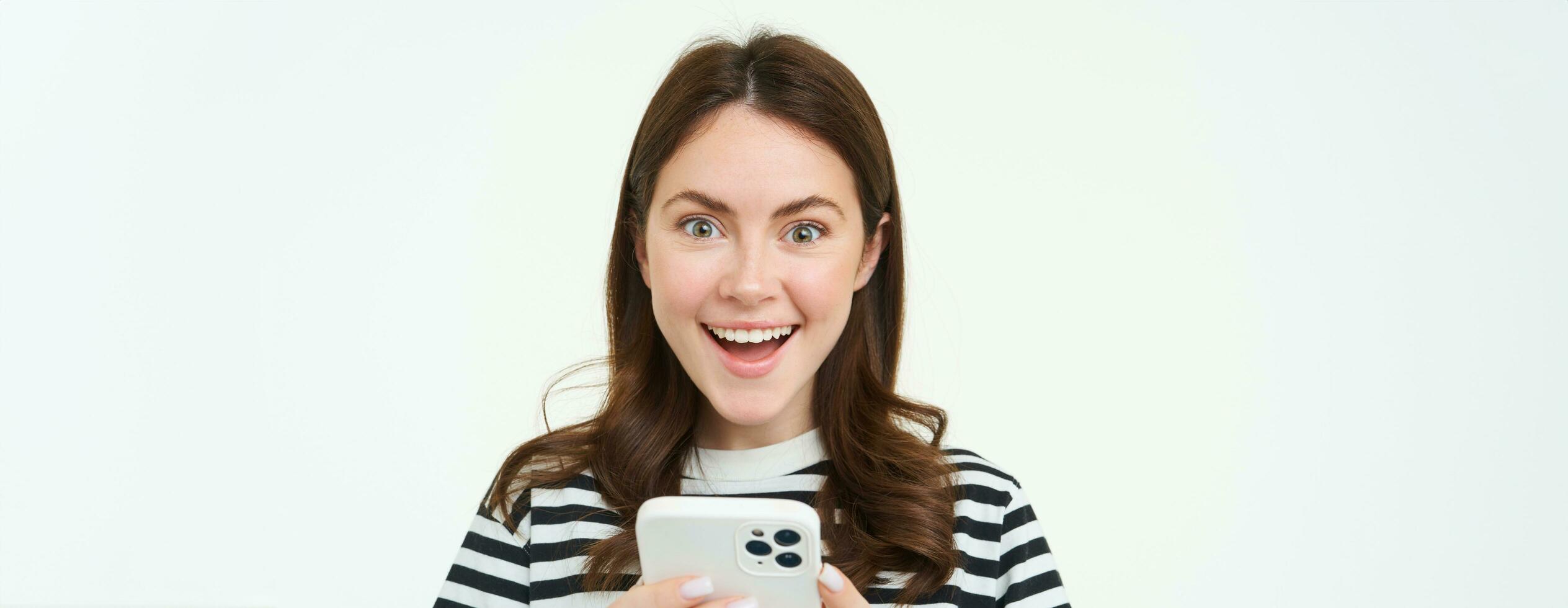 Portrait of excited brunette girl, reacting to amazing promo on smartphone, holding mobile phone and smiling, isolated on white background photo