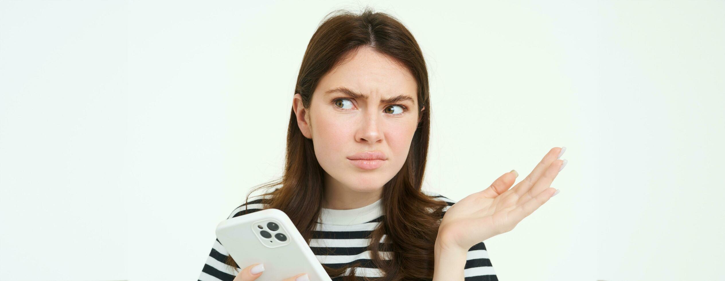 Portrait of angry, confused young woman shrugging shoulders while using mobile phone, holding smartphone with annoyed face expression, frowning, standing over white background photo