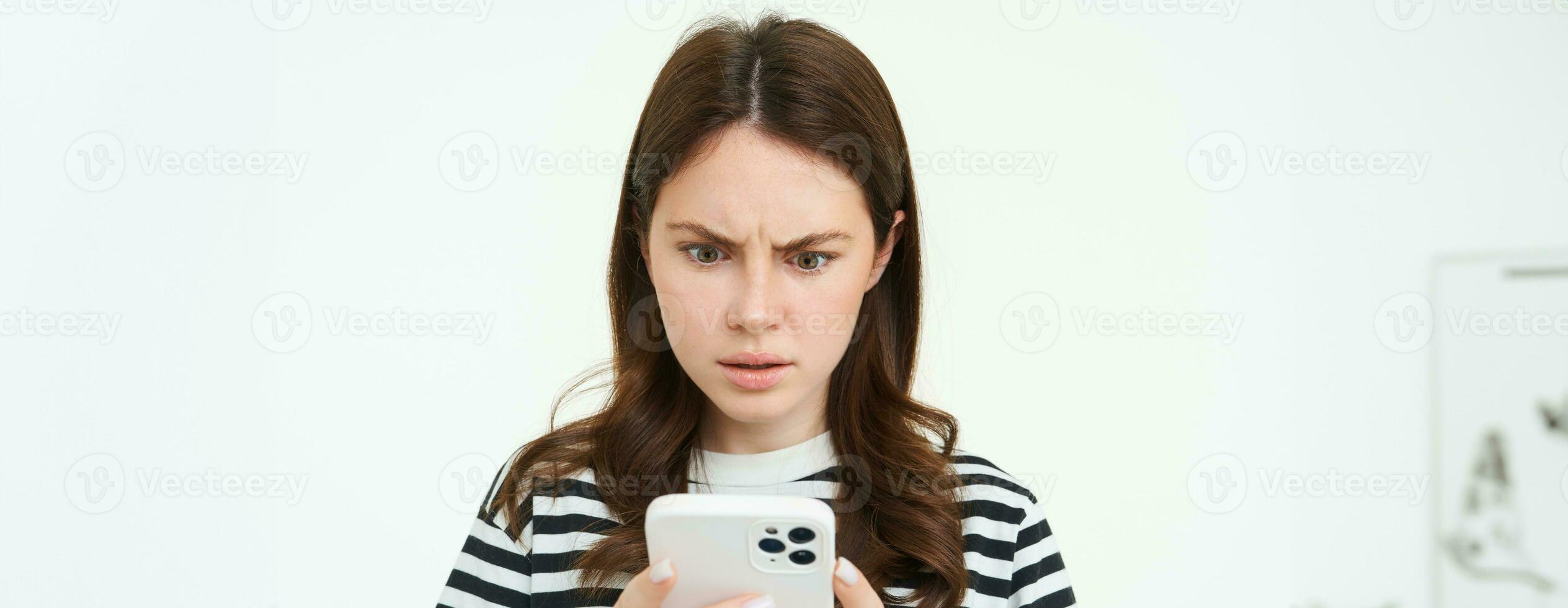 mujer mirando confuso y conmocionado a teléfono inteligente pantalla, ceñudo, leyendo molesto, concerniente a Noticias en móvil teléfono, aislado en blanco antecedentes foto