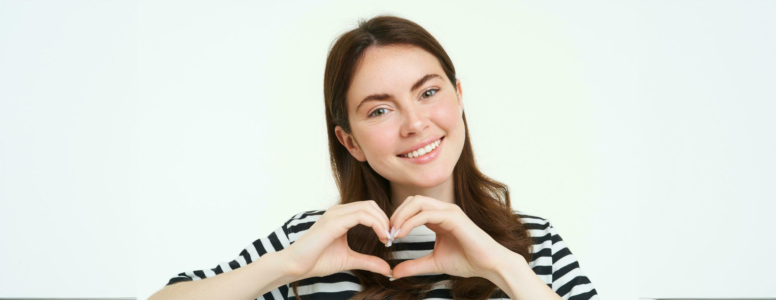 Portrait of smiling, happy girlfriend shows heart with hands and looking with care at camera, express her love or affection, like product, standing over white studio background photo