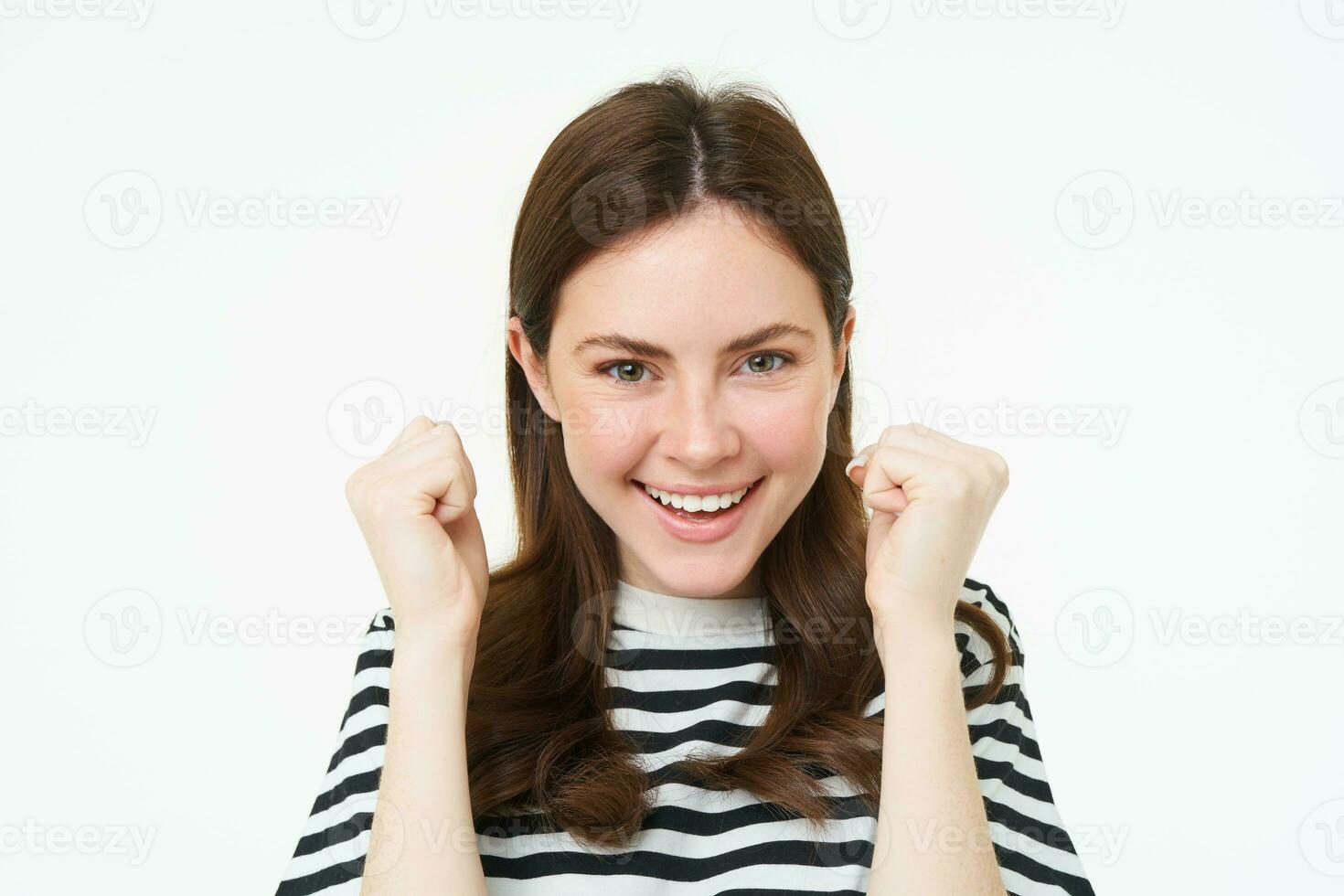 Image of young woman looking with pleased, satisfied face at camera, winning, shaking hands in triumph, celebrating victory, achievement, standing over white background photo