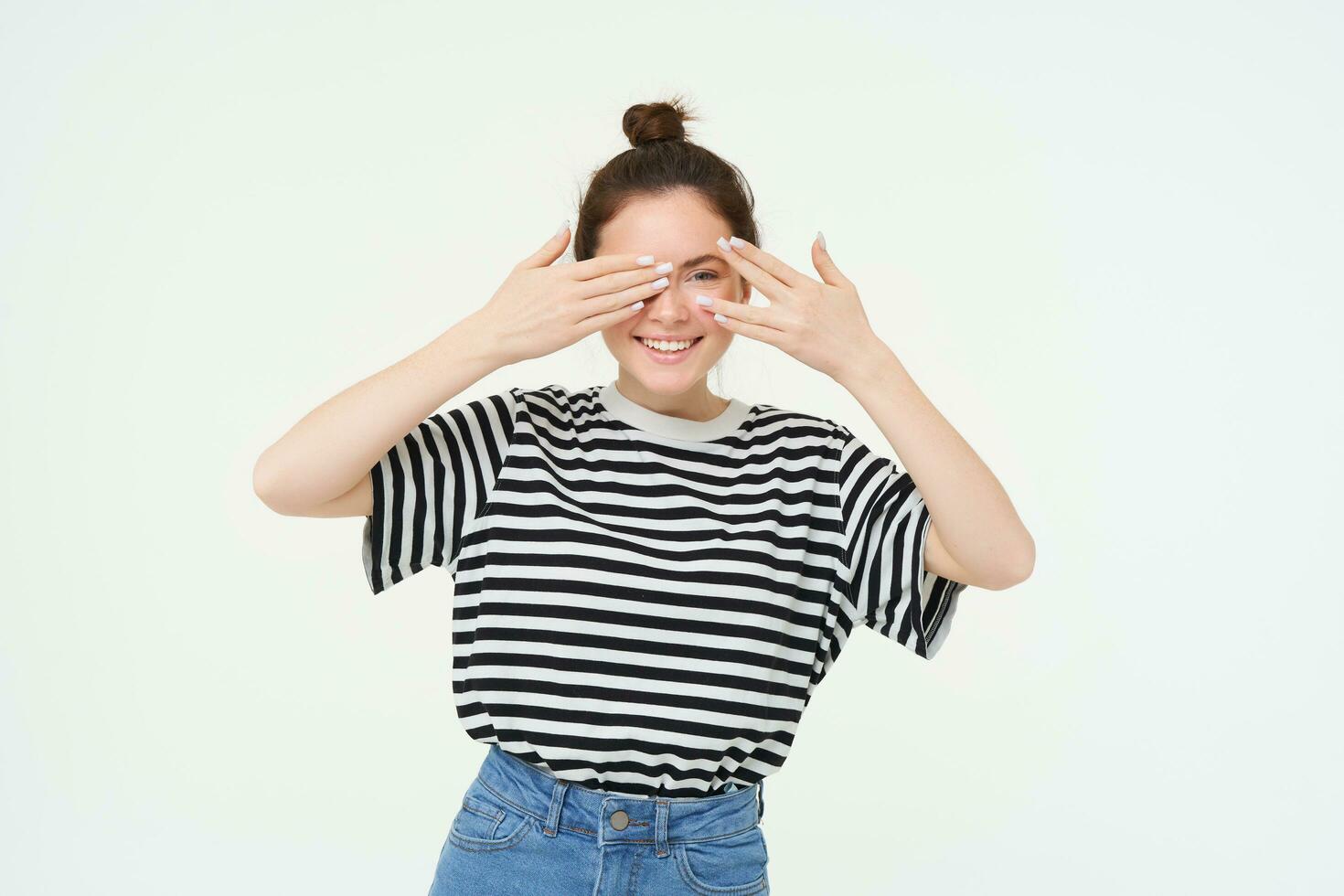 Portrait of girl waiting for surprise, covers eyes with hands, peeking through fingers, stands over white background photo