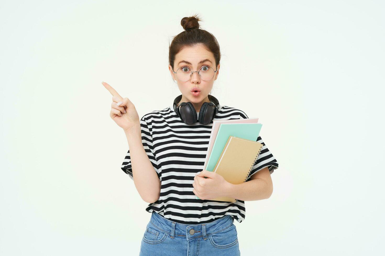 Portrait of girl with surprised face, interested in advertisement on the left, pointing at banner with curious face, wearing glasses, holding documents and notebooks in hand, white background photo