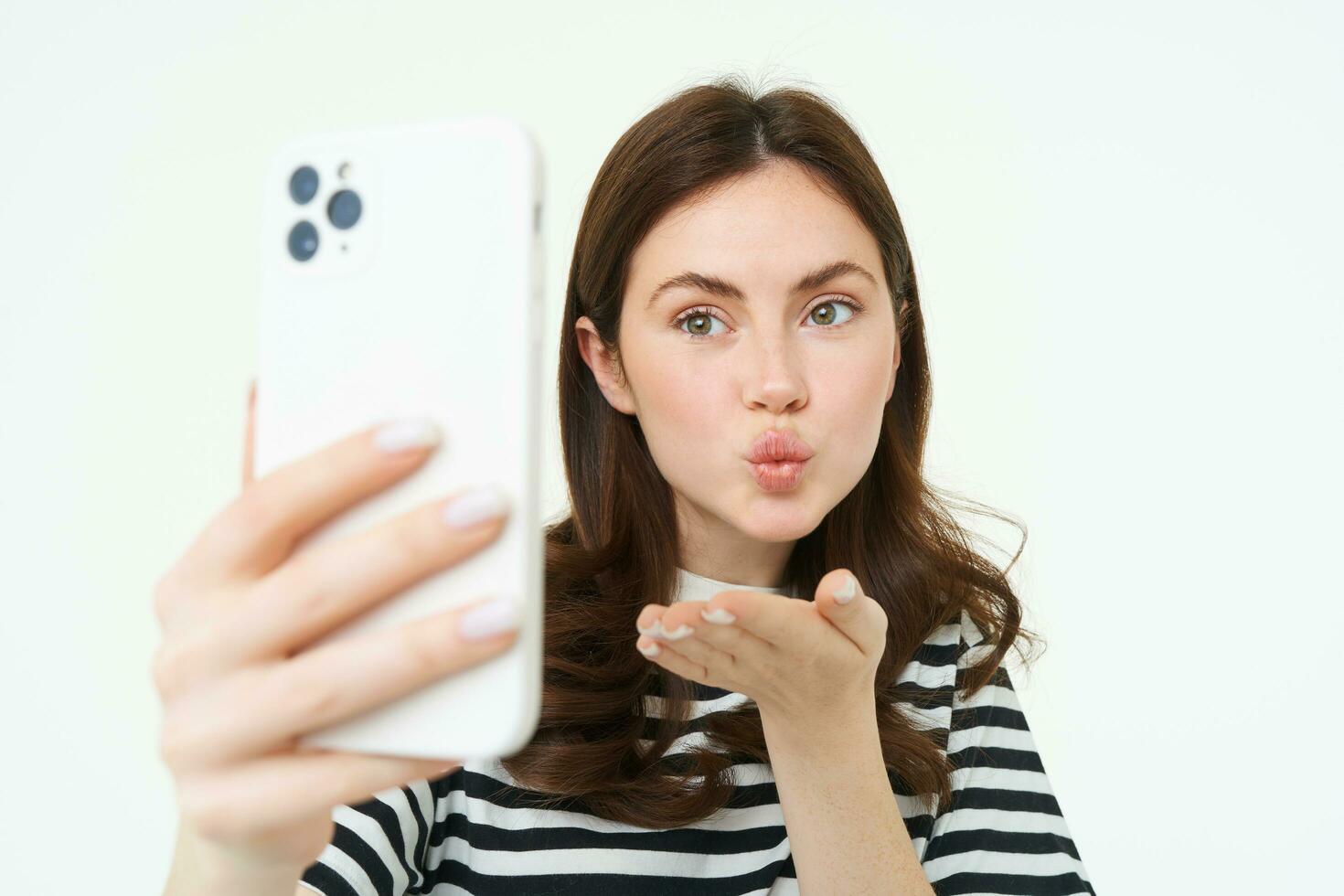 Portrait of young european woman taking selfie on smartphone, holding white mobile phone and posing for photos, isolated against white background photo
