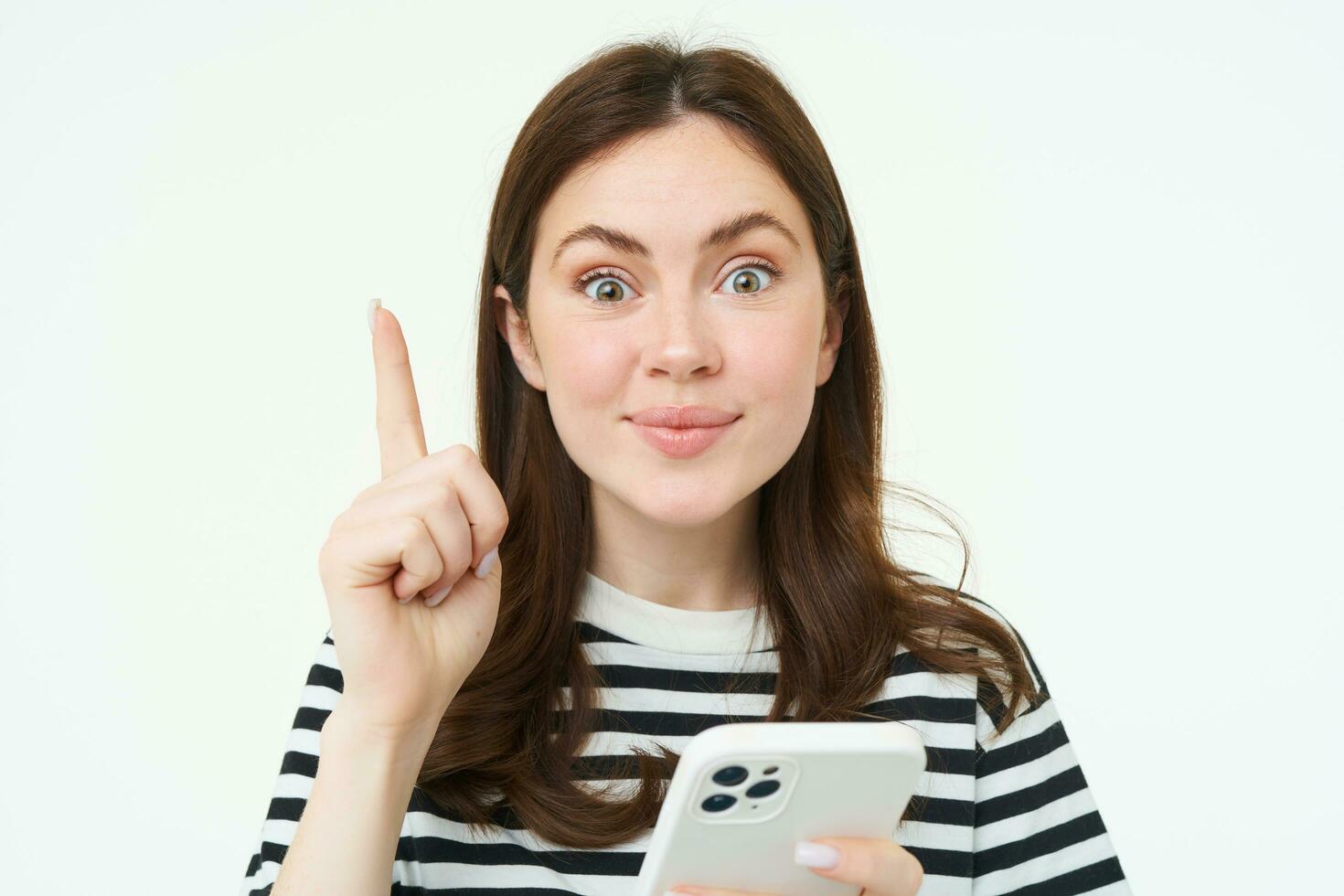 Portrait of beautiful young woman, has an idea, suggestion, shows one finger and holds smartphone, points up, white background photo