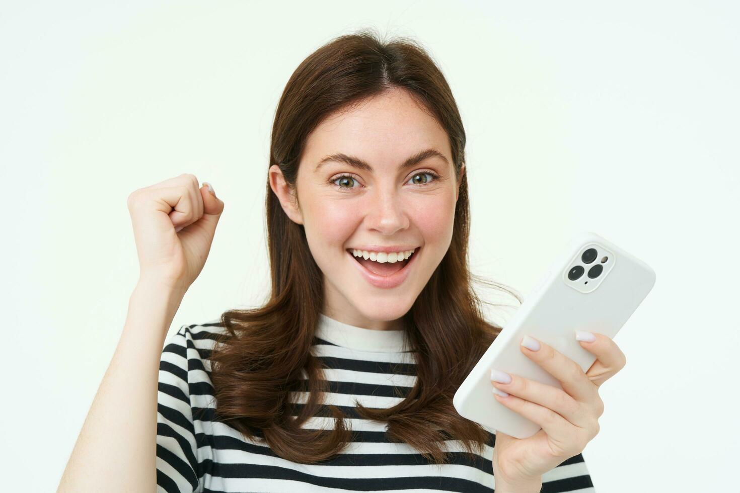 Enthusiastic brunette girl winning, holding smartphone and cheering, celebrating great news, standing with excited face expression, white background photo