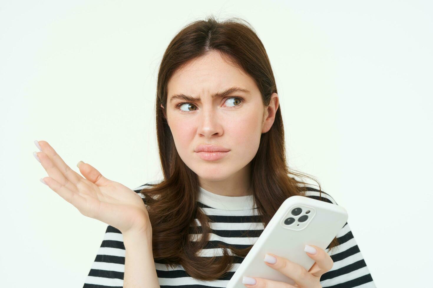 Portrait of angry, confused young woman shrugging shoulders while using mobile phone, holding smartphone with annoyed face expression, frowning, standing over white background photo