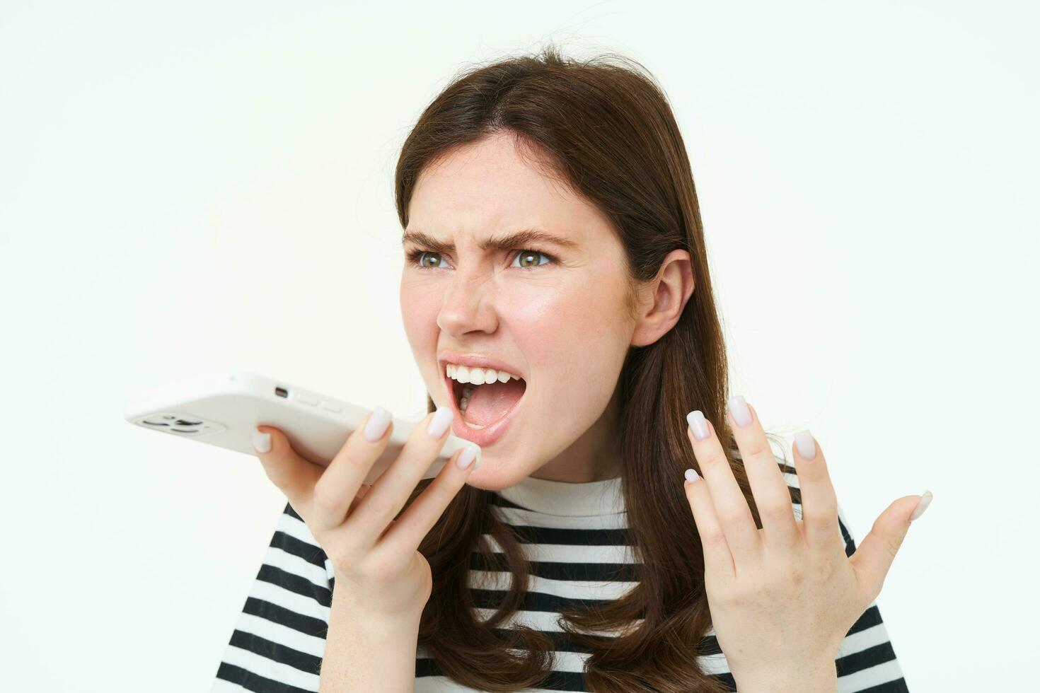 Portrait of angry woman shouting at smartphone speakerphone, recording voice message with annoyed face expression, screaming at phone, white background photo