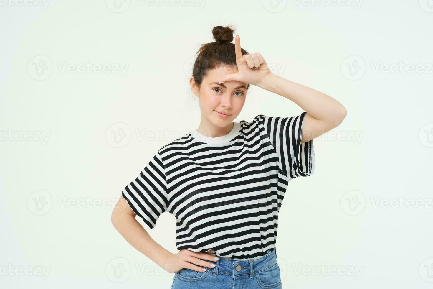 Portrait of brunette woman shows l letter on forehead, mocking someone, calling person a loser, standing over white background photo