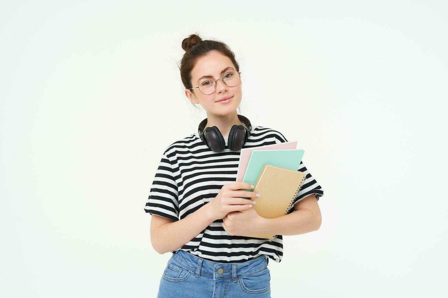 Student and education concept. Young woman with books, notes and pen standing over white background, college girl with headphones over neck posing in studio photo