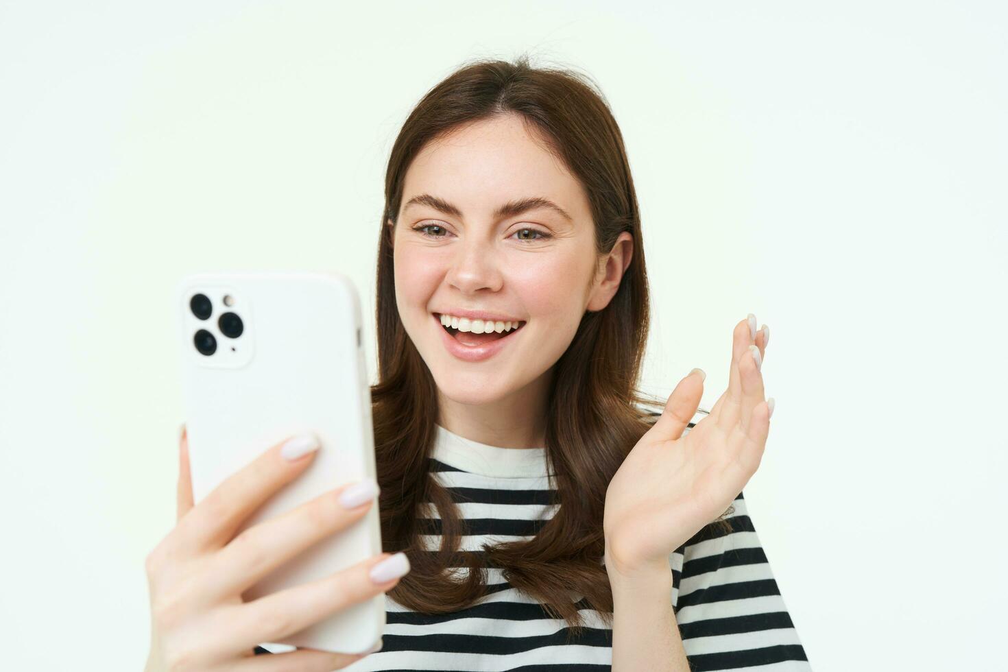 Portrait of young modern girl connects to video chats, talks with friend online using smartphone app, waves at mobile phone camera, white background photo