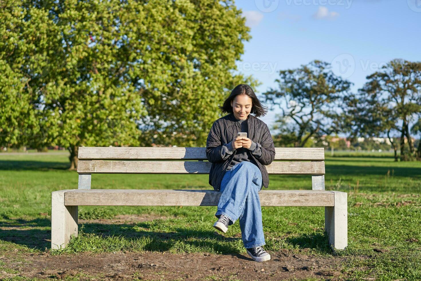 Young woman sitting alone on bench in park, using mobile phone, looking at screen, texting, sending messages in app chat photo