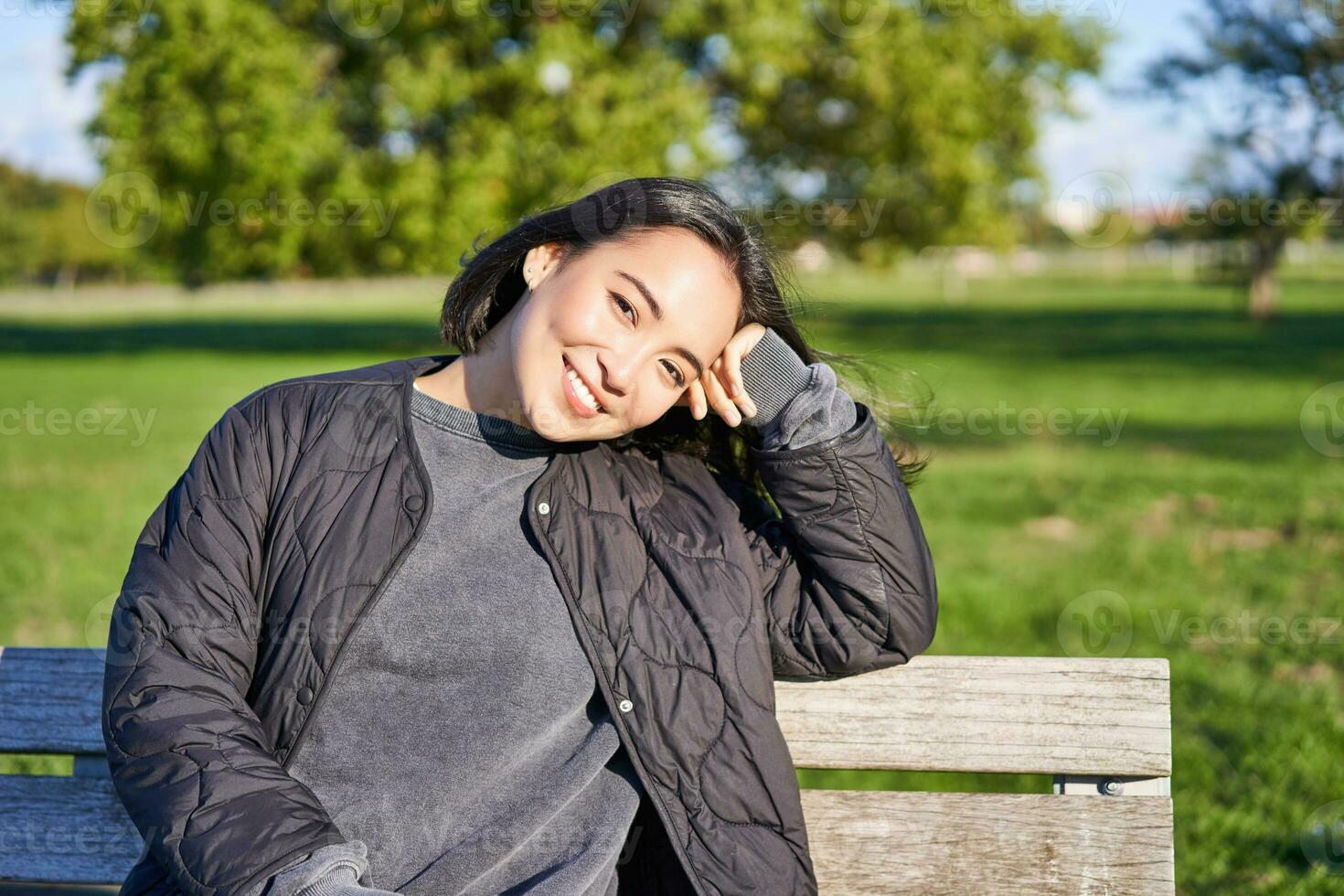 Beauty portrait of smiling asian girl with dark short hair, sitting on bench in park and gazing at camera photo