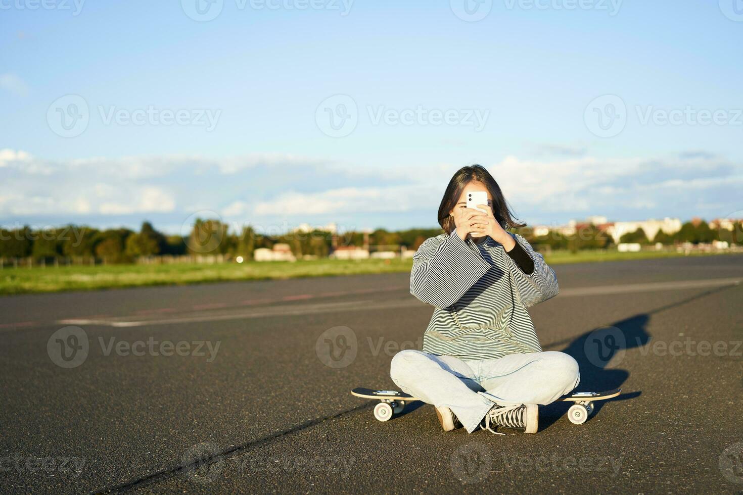 Hipster teen girl sitting on her skateboard, taking photos on smartphone. Asian woman skater sits on longboard and photographing on mobile phone