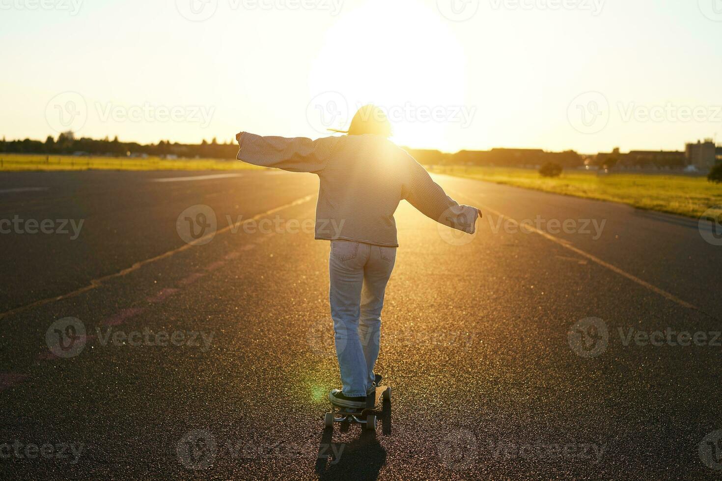 Rear view photo of young girl riding skateboard towards sunlight. Happy young woman on her cruiser, skating on longboard