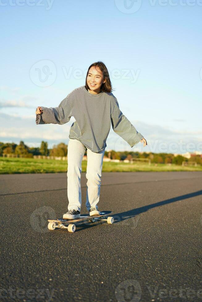 linda asiático niña montando patineta, Patinaje en la carretera y sonriente. patinador en crucero longboard disfrutando al aire libre en soleado día foto