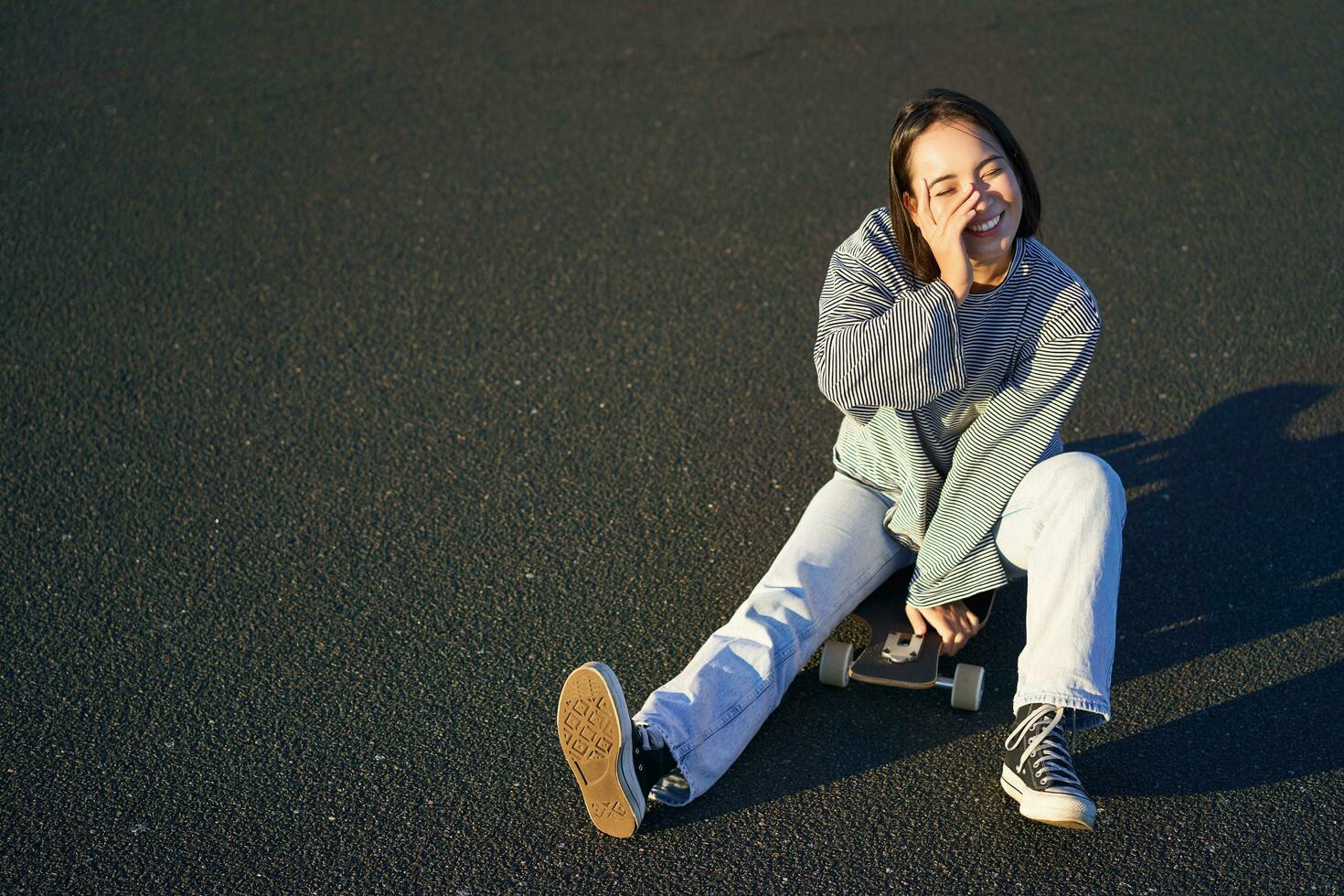 Happy beautiful korean teen girl sits on her skateboard, cruising on longboard, wearing casual clothes photo