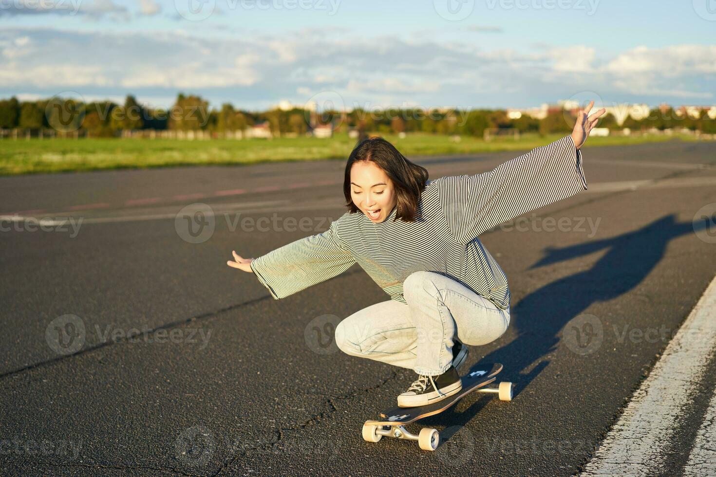 Freedom and happiness. Happy asian girl riding her longboard on an empty sunny road, laughing and smiling, skateboarding photo