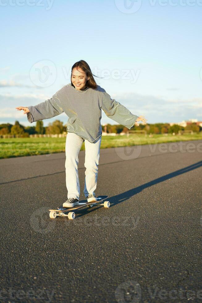 linda asiático niña montando patineta, Patinaje en la carretera y sonriente. patinador en crucero longboard disfrutando al aire libre en soleado día foto