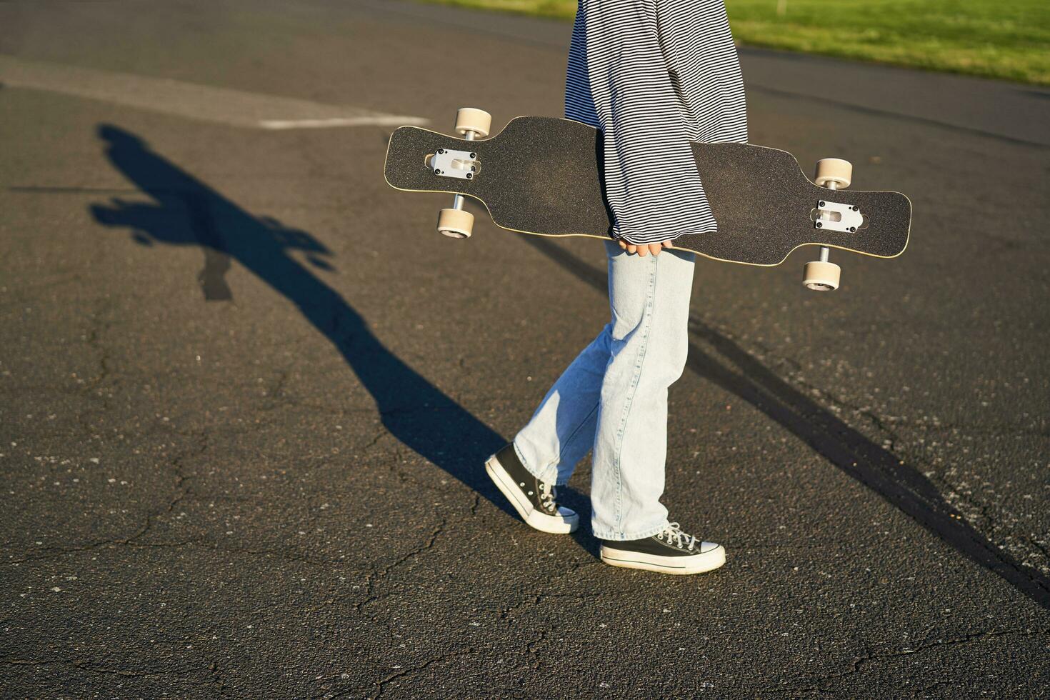 Cropped shot of teen girl body, holding cruiser longboard in hand, walking in sneakers on road in jeans and sweatshirt. Young woman skater with skateboard photo