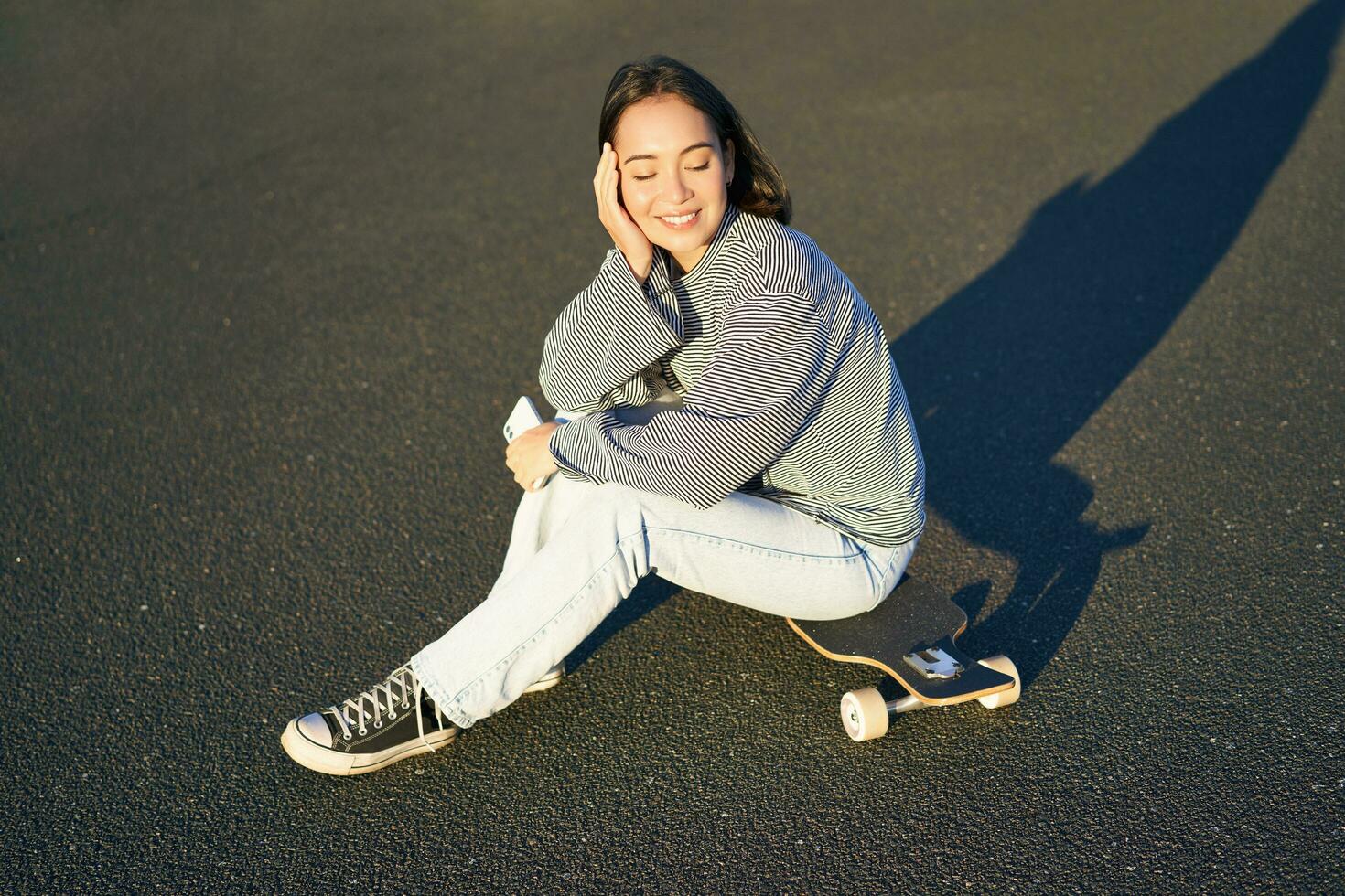 Portrait of asian woman sitting on skateboard, skating on her cruiser longboard, using smartphone app photo