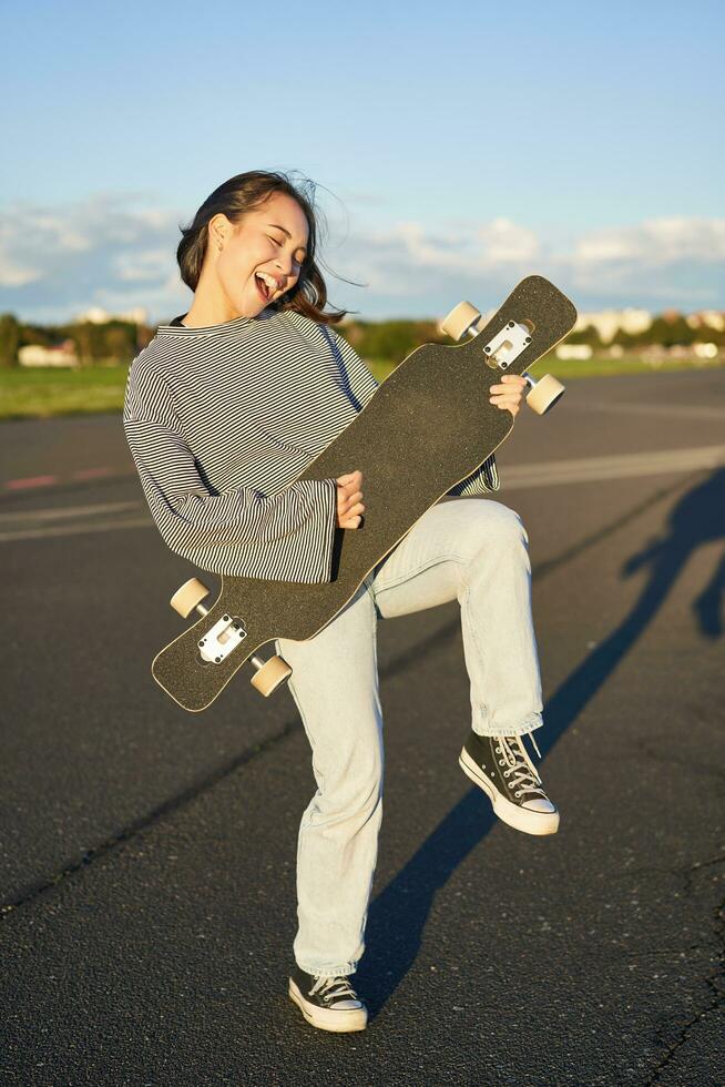 gracioso asiático niña disfrutando Patinaje, participación patineta me gusta guitarra y sombra jugando, teniendo divertido al aire libre foto