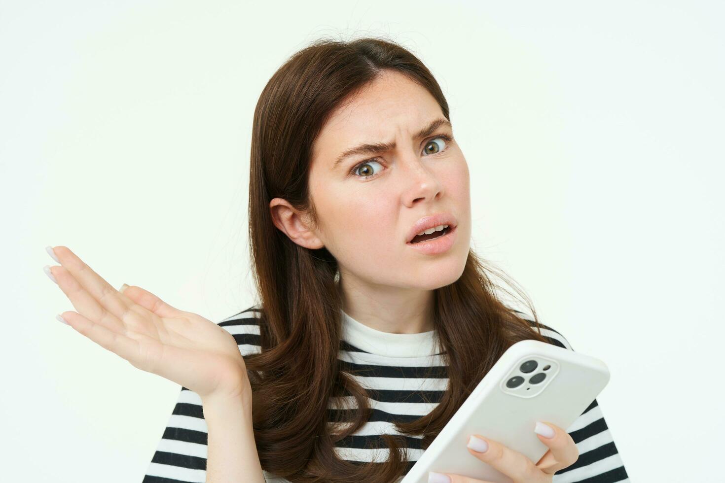 Portrait of woman shrugging, holding smartphone and looking confused at camera, isolated on white background photo