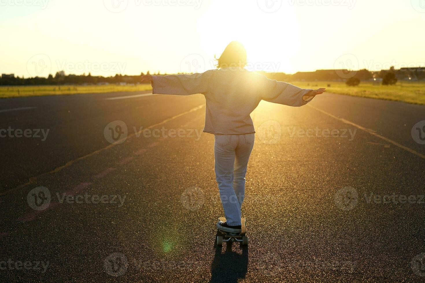 Carefree teen skater girl riding her skateboard with hands spread sideways, skating alone towards the sun photo