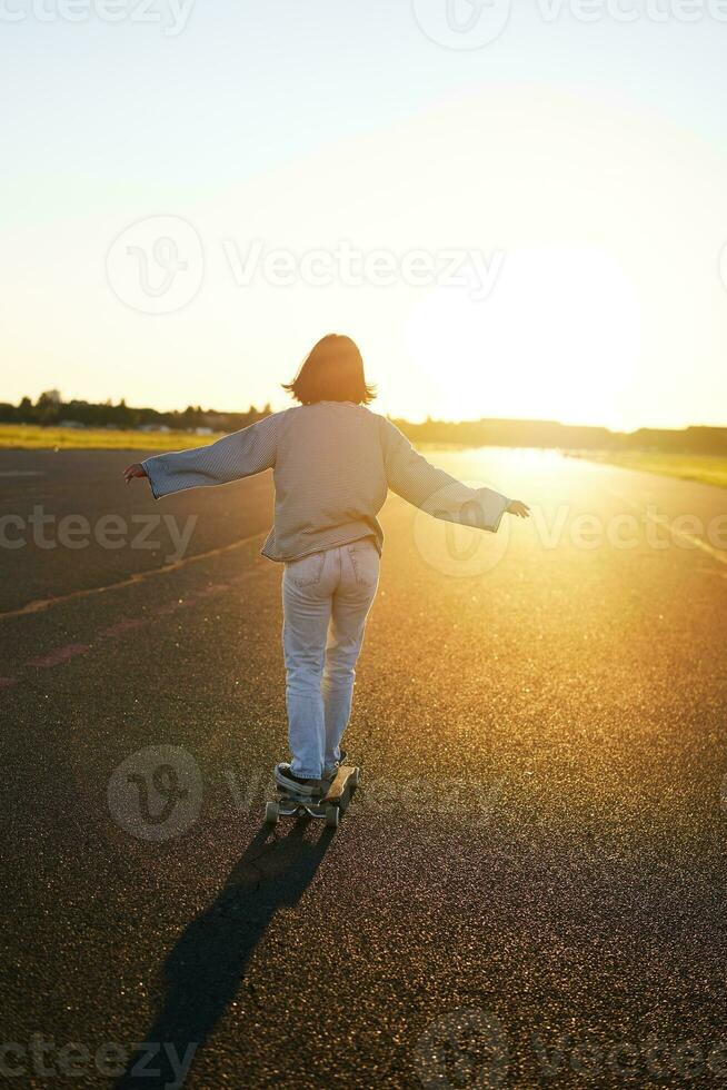 Rear view photo of young girl riding skateboard towards sunlight. Happy young woman on her cruiser, skating on longboard