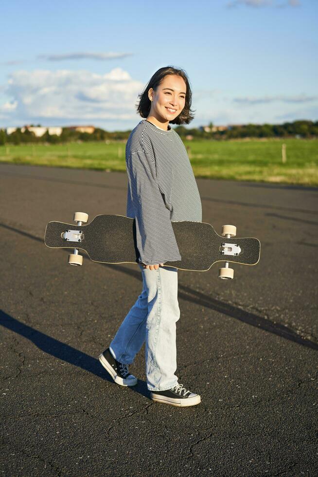 vertical Disparo de despreocupado asiático niña con longboard joven mujer patinador participación crucero en su espalda y caminando en camino, patinar foto