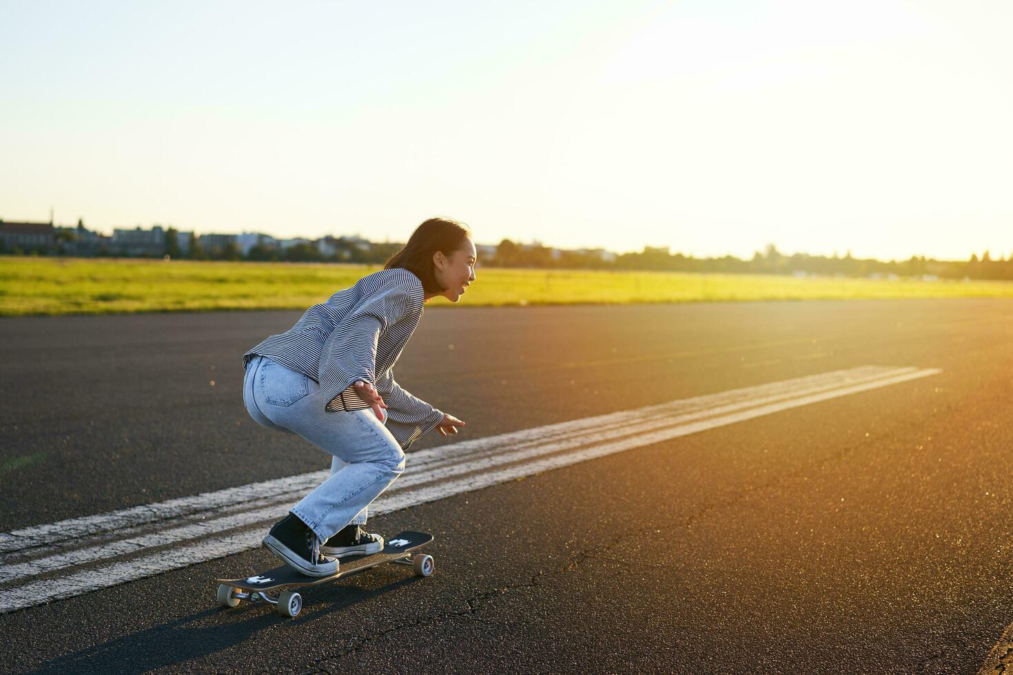 Side view of beautiful asian girl on skateboard, riding her cruiser towards the sun on an empty road. Happy young skater enjoying sunny day on her skate photo