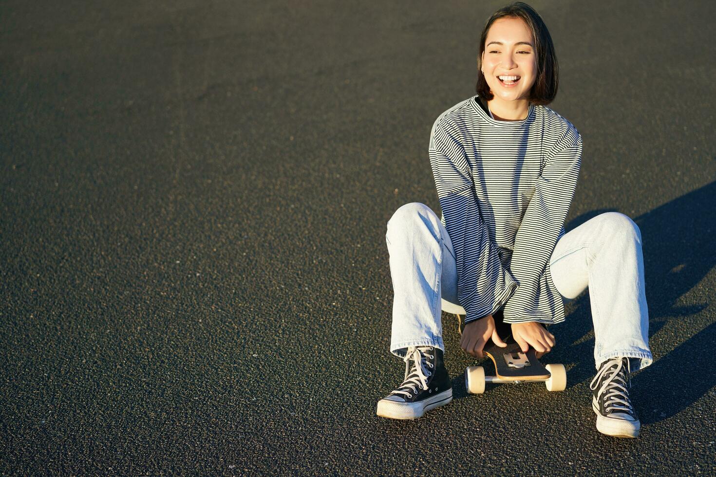 libertad y felicidad. linda sonriente asiático chica, se sienta en patineta en soleado primavera día. contento riendo patinador disfrutando crucero en longboard foto