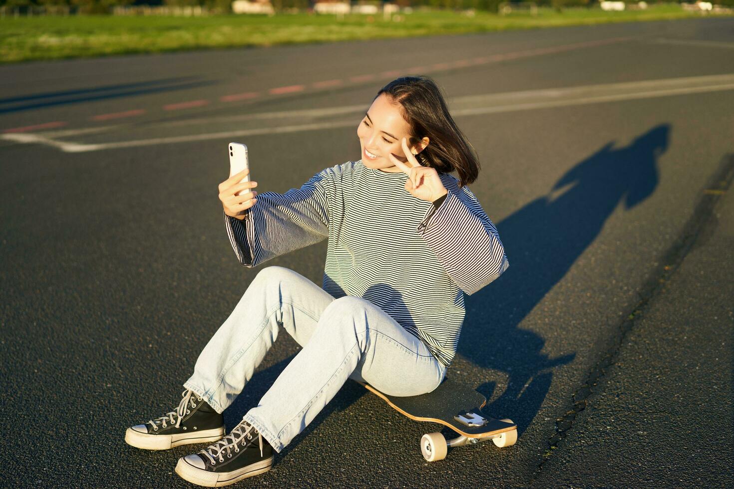 Happy asian girl sits on skateboard, takes selfie with longboard, makes cute faces, sunny day outdoors photo