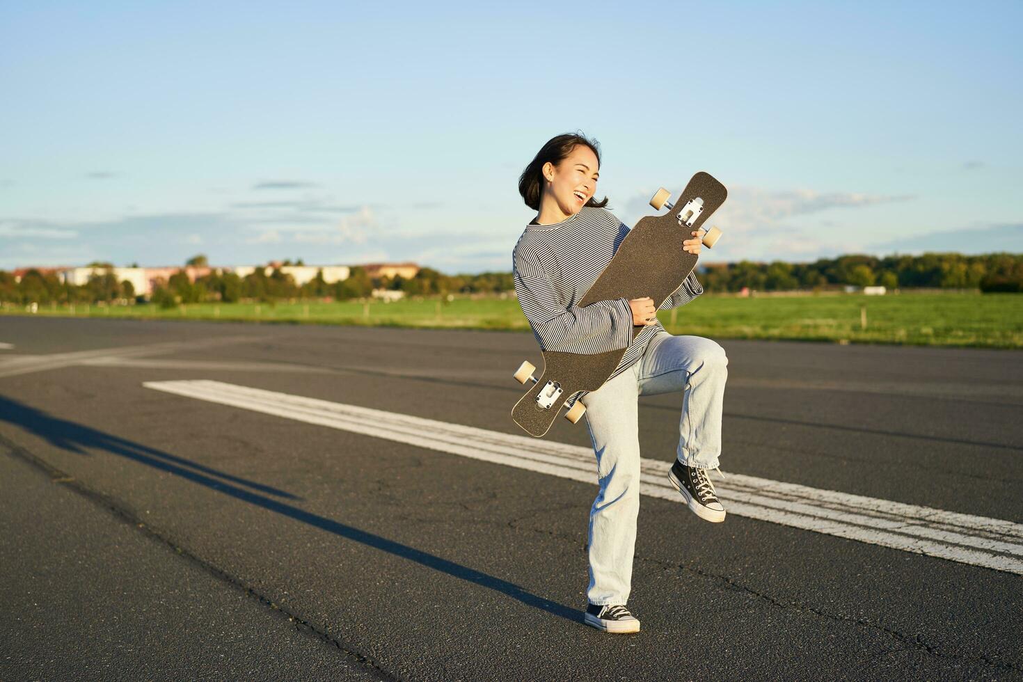 contento morena asiático niña teniendo divertido en calle con longboard, Patinaje, utilizando patineta como guitarra, disfrutando patinar foto