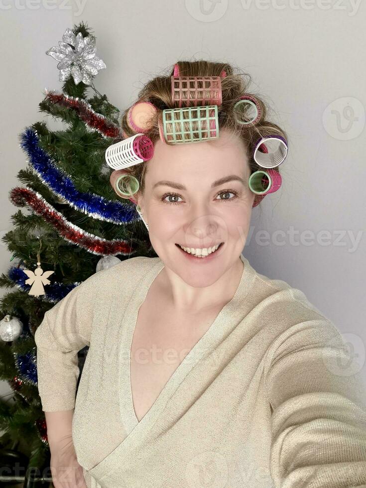 Young girl with fair skin in light sweater with curlers on her hair on the background of a christmas tree taking a selfie photo